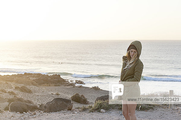 Blondes Mädchen am Strand mit Jacke und Mütze bei Sonnenuntergang
