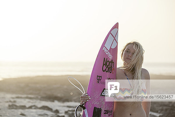 Blondes Surfermädchen steht allein am Strand bei Sonnenuntergang mit ihrem Surfbrett