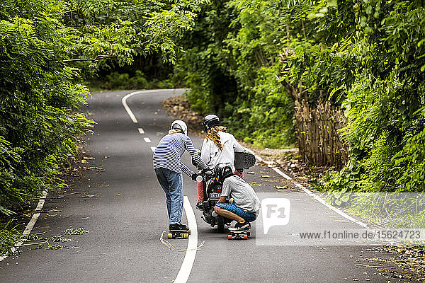 Zwei Longboarder tragen ein Motorrad hinter sich her  das einen dritten Skateboarder trägt