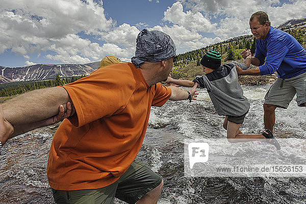 Two men help a boy as he leaps from stone to stone across a raging stream  a tributary of Yellowstone Creek  in the alpine tundra below Kings Peak  on the fourth day of Troop 693's six day backpacking trip in the High Uintas Wilderness Area  Uintas Range  Utah