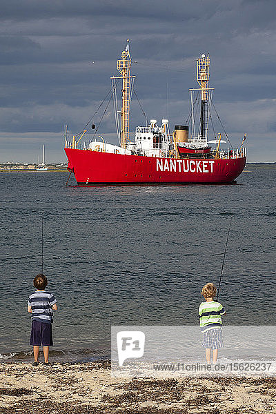Zwei Jungen angeln am Brant Point-Leuchtturm von Nantucket. Im Hintergrund ist das Feuerschiff von Nantucket zu sehen  das auch dazu beiträgt  Boote sicher in den und aus dem Hafen von Nantucket zu steuern.
