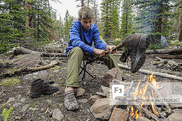 A boy dries his wet boots over a fire at camp  after a rainy day of hiking  the first of Troop 693's six day backpack trip through the High Uintas Wilderness Area  Uintas Range  Utah