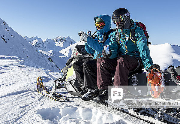 Snowboarder sitzen auf einer Maschine und genießen ein Bier mit Blick auf ferne Berge