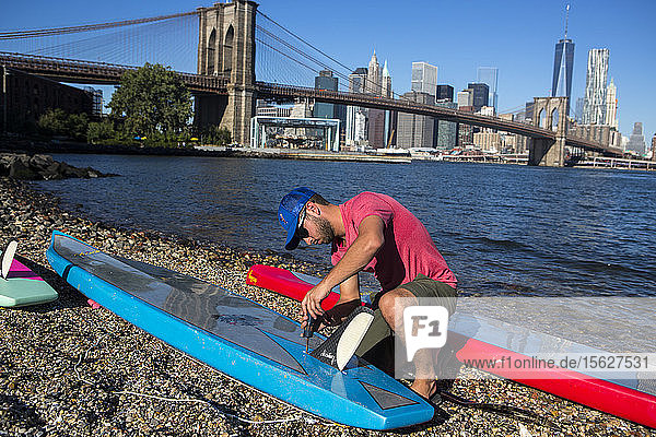 Man crouching on riverbank preparingï¾ paddleboardsï¾ with Brooklyn Bridge in background  New York City  New York  USA