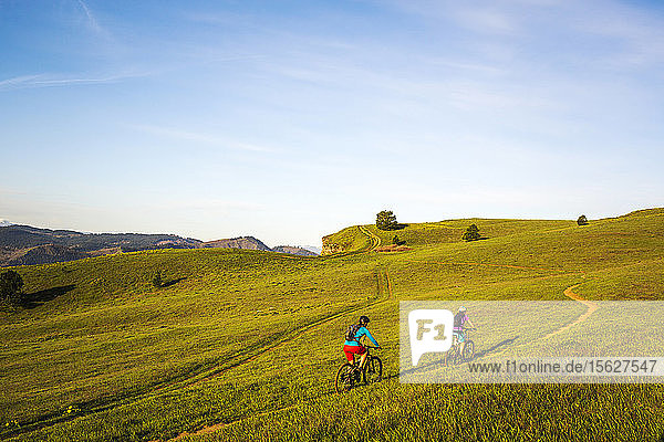 Two young women ride mountain bikes up a single-track trail through an open meadow.
