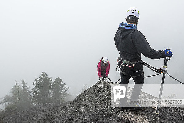 Ein Mann wartet und schaut zurück  während eine Frau die Metallsprossen eines Klettersteigs in Squamish  British Columbia  hinaufklettert.