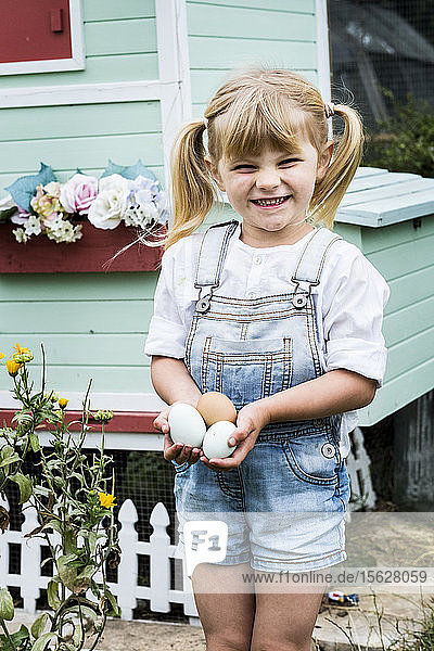 Blond girl standing in a garden in front of hen house  holding fresh eggs.