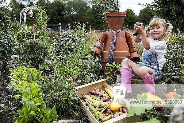 Lächelndes blondes Mädchen sitzt im Garten neben der Terrakotta-Vogelscheuche und pflückt frisches Gemüse.