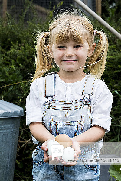 Blondes Mädchen steht in einem Garten  hält frische Eier in der Hand und lächelt in die Kamera.