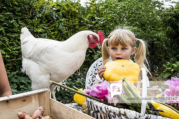 Blondes Mädchen mit gelbem Kürbis und weißem Huhn in einem Garten.