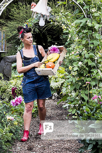 Frau  die in einem Garten spazieren geht und eine Holzkiste mit frischem Gemüse und Blumen trägt.