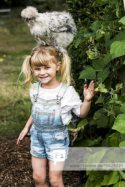 Lächelndes blondes Mädchen  das in einem Garten steht  mit einem flauschigen grauen Huhn auf dem Kopf.