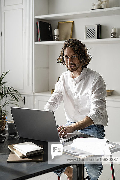 Porträt eines selbstbewussten Mannes  der zu Hause am Tisch sitzt und seinen Laptop benutzt