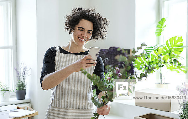 Lächelnde junge Frau beim Fotografieren von Blumen in einem kleinen Geschäft mit einem Smartphone
