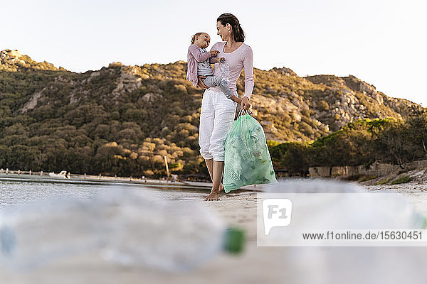 Frau am Strand mit kleiner Tochter auf dem Arm sammelt leere Plastikflaschen