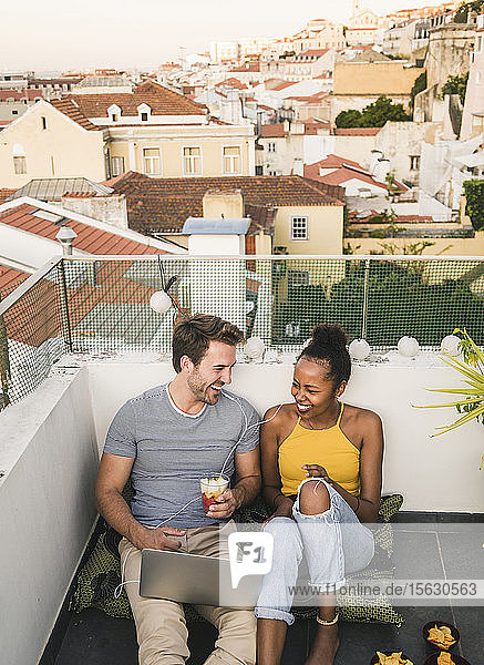 Glückliches junges Paar mit Laptop und Kopfhörern sitzt abends auf dem Dach  Lissabon  Portugal