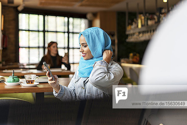 Junge Frau trägt einen türkisfarbenen Hijab und benutzt ein Smartphone in einem Cafe