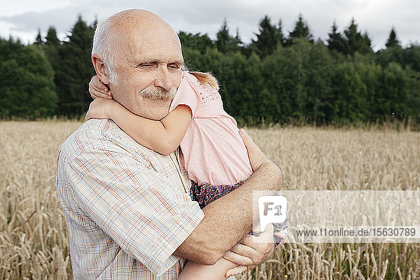 Porträt eines älteren Mannes in einem Haferfeld  der seine Enkelin auf dem Arm trägt