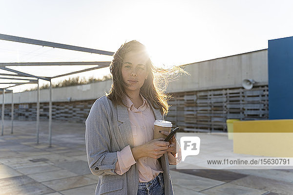 Porträt einer jungen Frau mit Kaffee zum Mitnehmen und Mobiltelefon bei Gegenlicht