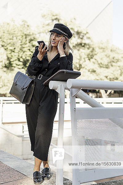 Young blond businesswoman wearing black hat and using smartphone