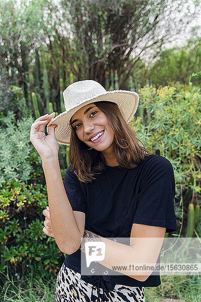 Porträt einer lächelnden jungen Frau mit Tätowierung mit Sommerhut in der Natur