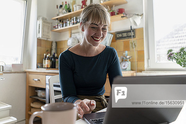 Porträt einer glücklichen Frau  die mit einem Laptop in der Küche sitzt