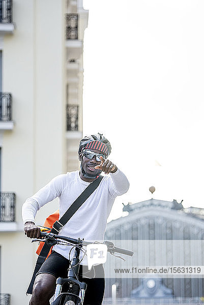 Stilvoller junger Mann mit Fahrrad und Kuriertasche in der Stadt  der mit dem Finger zeigt
