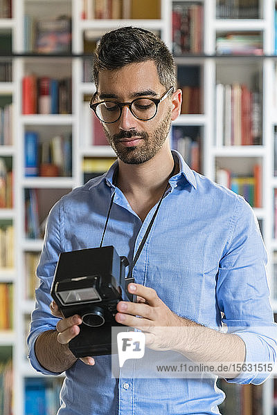 Porträt eines jungen Mannes  der vor einem Bücherregal steht und auf eine Sofortbildkamera schaut