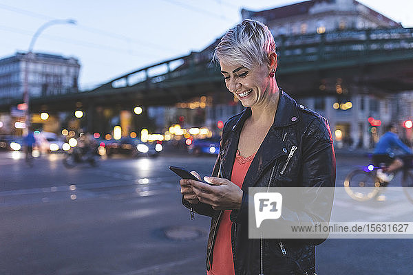 Porträt einer lachenden blonden Frau  die abends mit dem Handy am Straßenrand steht  Berlin  Deutschland