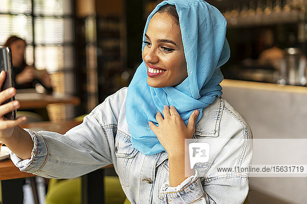 Junge Frau trägt einen türkisfarbenen Hijab und benutzt ein Smartphone in einem Cafe