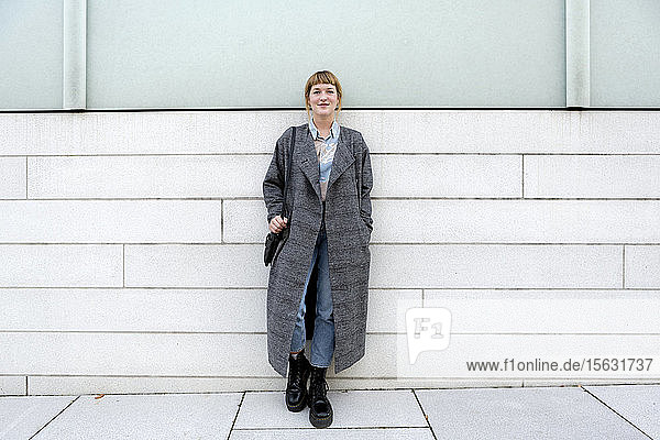 Portrait of strawberry blonde young woman wearing grey coat leaning against wall