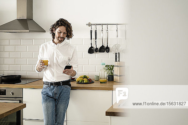 Man holding glass of orange juice and cell phone in kitchen at home