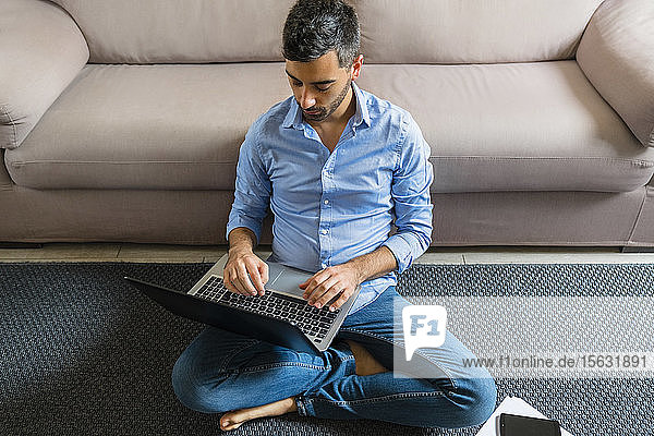 Junger Mann sitzt zu Hause mit Laptop auf dem Boden