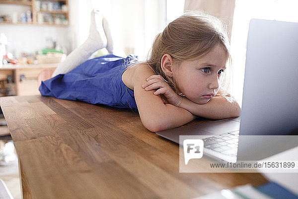 Portrait of sad little girl lying on kitchen table at home looking at laptop