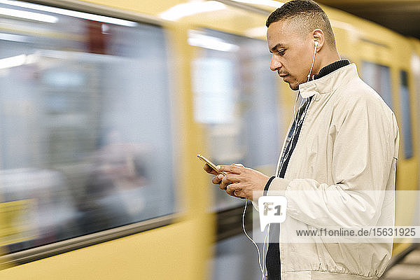 Man standing at underground station platform using earphones and cell phone  Berlin  Germany