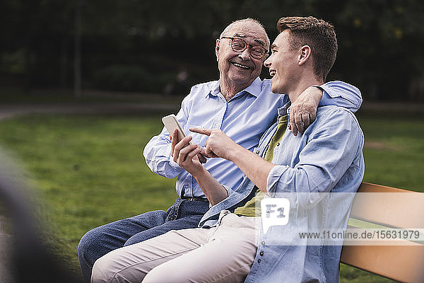 Älterer Mann und Enkel mit Smartphone sitzen zusammen auf einer Parkbank und amüsieren sich