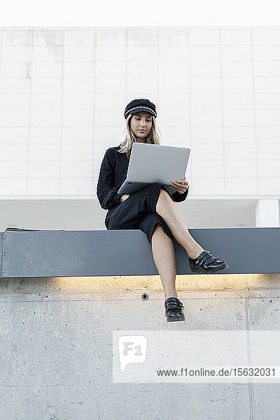 Young blond businesswoman wearing black sailor's cap and using laptop  sitting on a wall