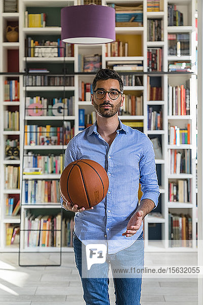Porträt eines jungen Mannes mit Basketball  der zu Hause vor einem Bücherregal steht
