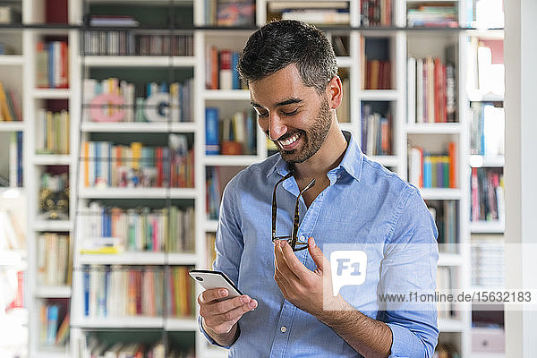 Porträt eines lächelnden jungen Mannes  der vor einem Bücherregal steht und auf sein Handy schaut