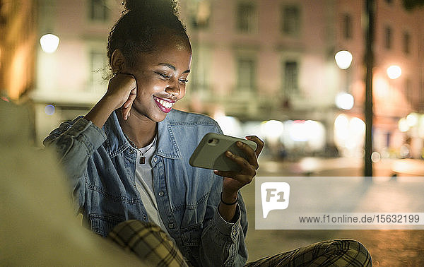 Porträt einer glücklichen jungen Frau  die ein Smartphone in der Stadt bei Nacht benutzt  Lissabon  Portugal