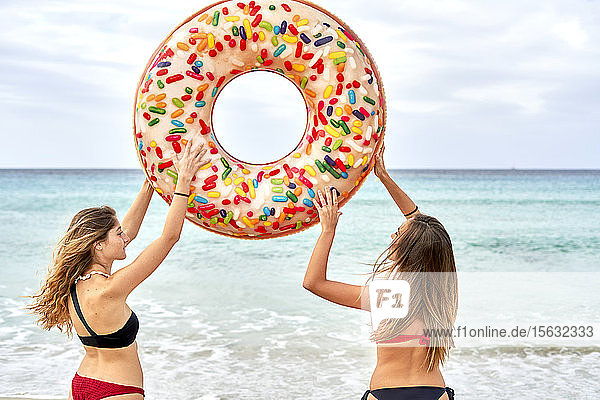 Zwei junge Frauen stehen vor dem Meer und halten einen großen aufblasbaren Ring in der Luft