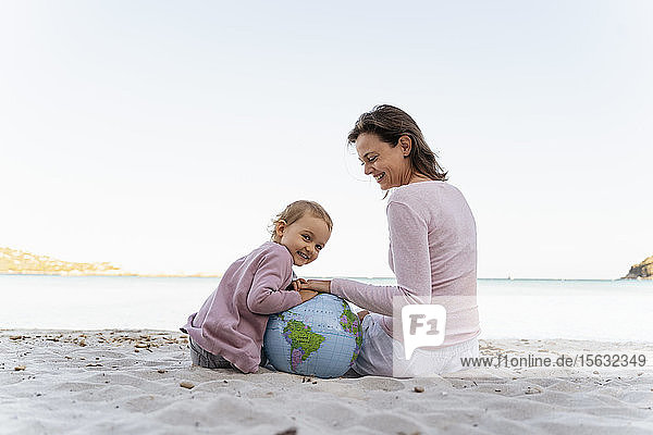 Porträt eines glücklichen kleinen Mädchens  das mit seiner Mutter am Strand sitzt und mit dem Earth Beachball spielt