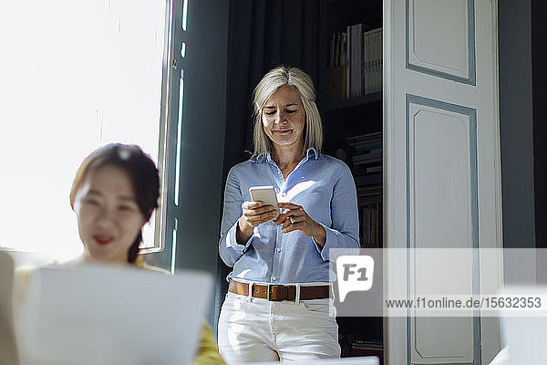 Eine reife Frau benutzt ein Smartphone im Hintergrund  während ein Kollege am Schreibtisch arbeitet