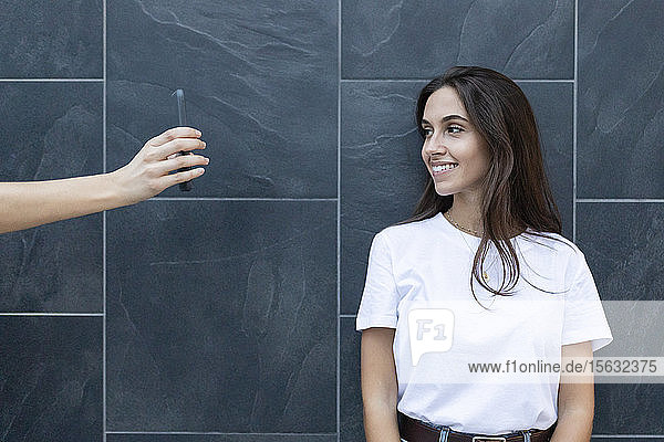 Junge lächelnde Frau schaut auf ein Smartphone  das ihr Freund in der Hand hält  mit einer grauen Wand im Hintergrund