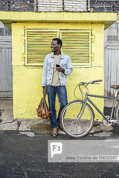 Junger Mann mit Smartphone  der vor dem gelben Verteilerkasten wartet