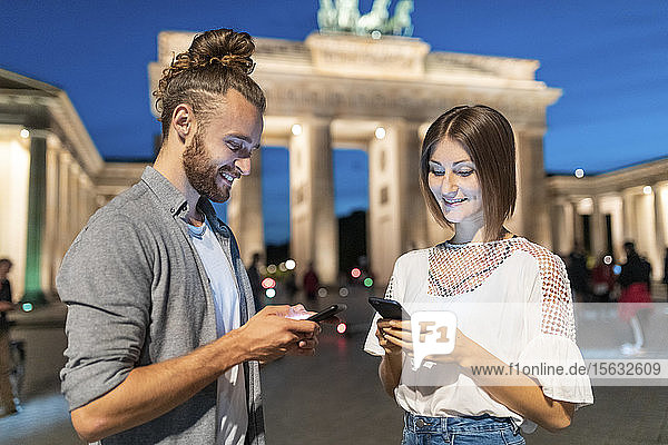 Glückliches Paar benutzt Smartphones am Brandenburger Tor zur blauen Stunde  Berlin  Deutschland