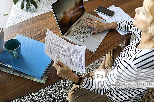 Reife Frau mit Dokumenten  die zu Hause einen Laptop benutzen