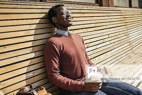 Junger Mann sitzt auf Holzbank  isst Hamburger  trinkt Kaffee