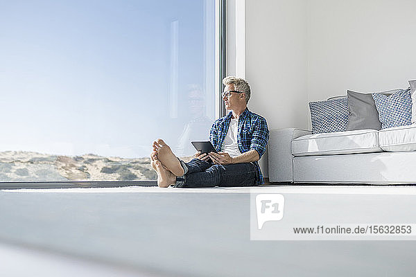 Lässiger Geschäftsmann sitzt in modernem Haus vor dem Fenster und hält Tablet