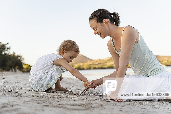 Mutter und Tochter zeichnen mit kleinen Stöckchen im Sand am Strand